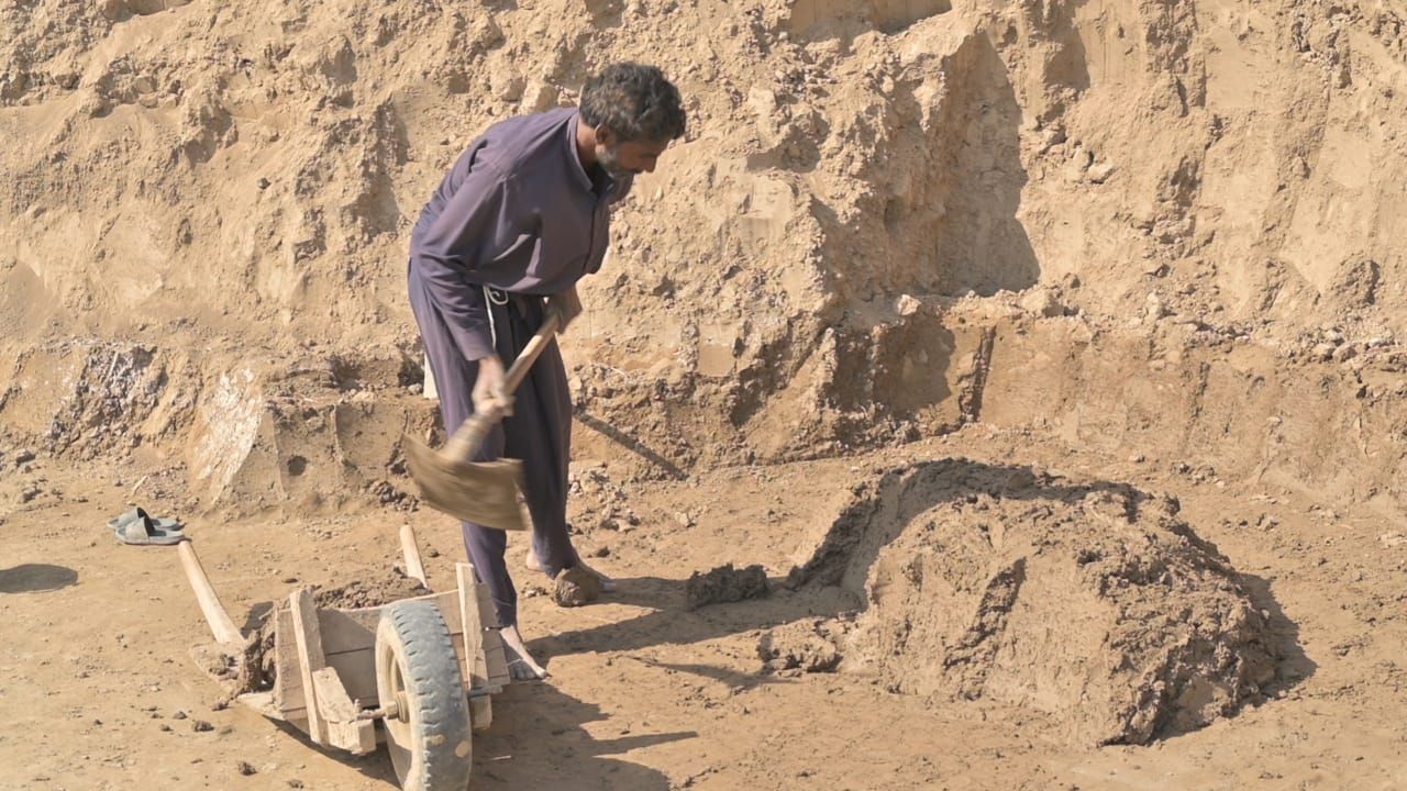 Man digging with a pickaxe in a sandy area, near a wooden cart.