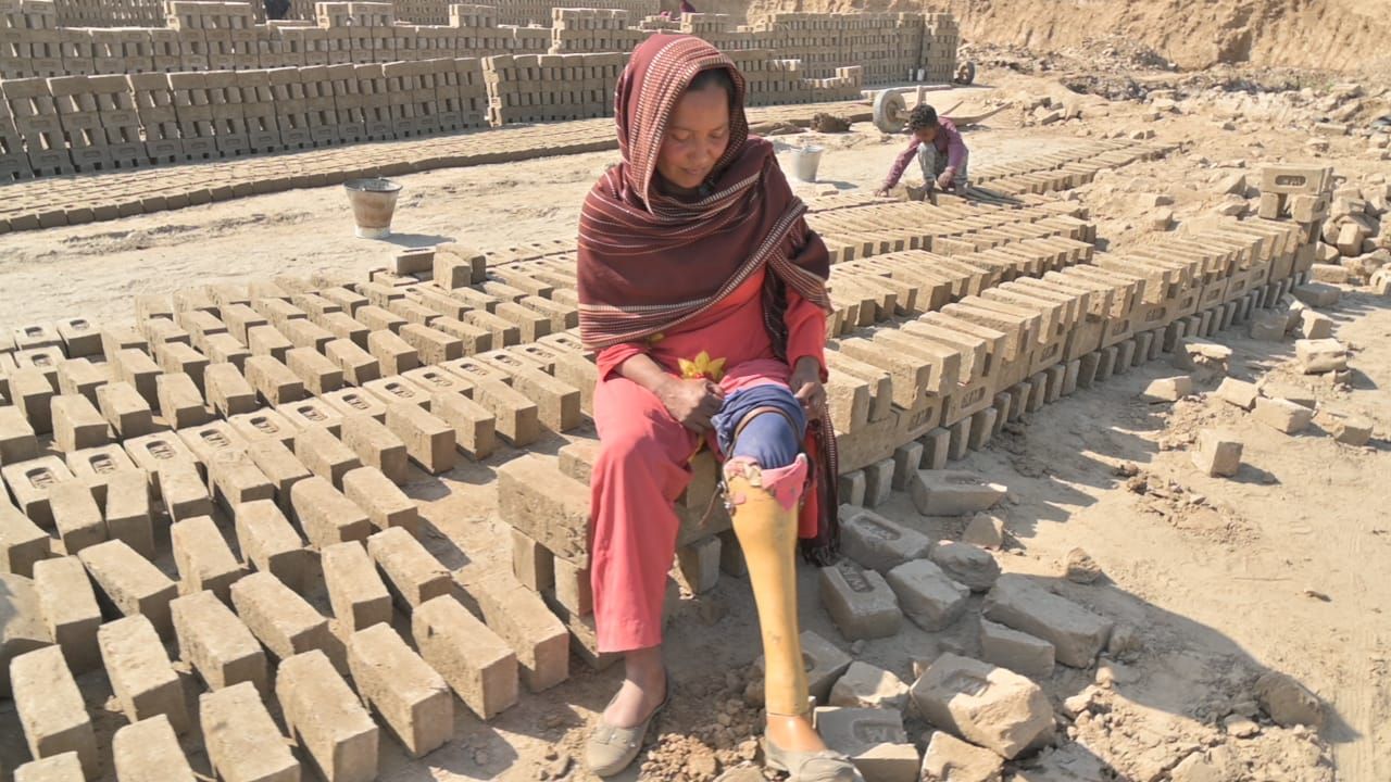 Woman with prosthetic leg sits among brick stacks at a brickyard, with a person working in the background.