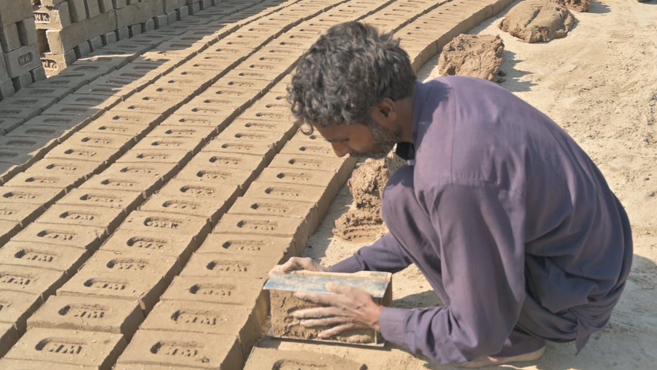 Man making bricks outdoors, squatting, arranging wet mud bricks in rows on dry ground.