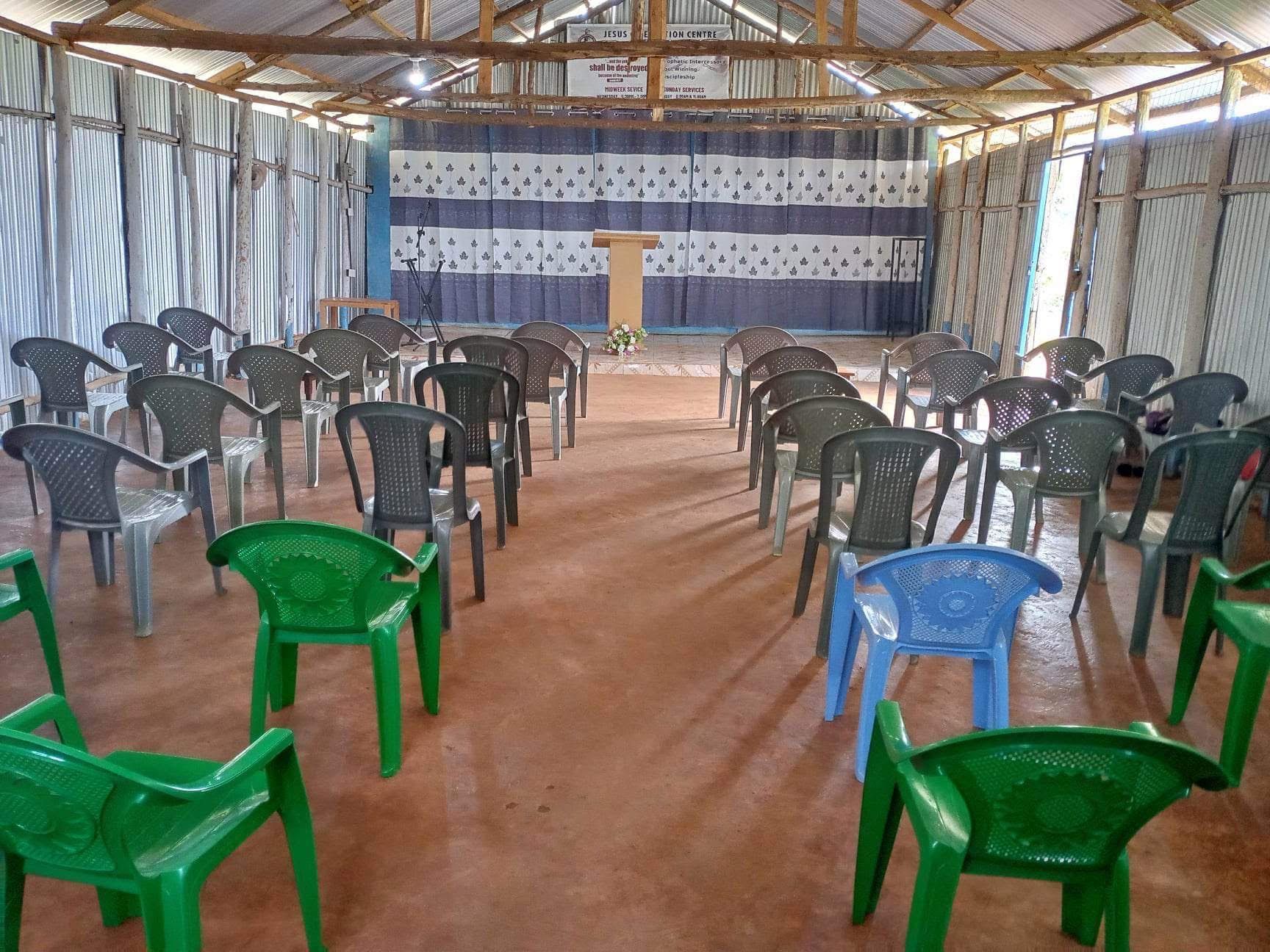 An empty church interior with rows of plastic chairs facing a wooden podium; blue and white curtains in the background.
