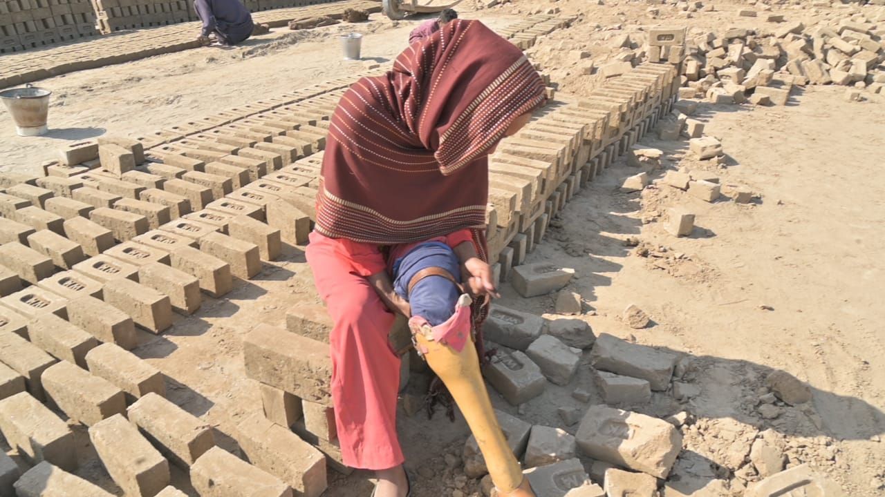 Woman working with bricks; brown head covering, pink pants, sunlight, outdoors.
