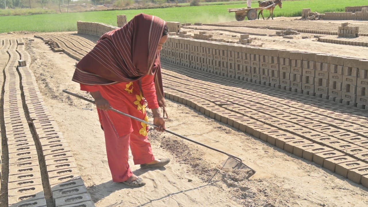 Woman in red pants and headscarf tending to rows of drying bricks outdoors.