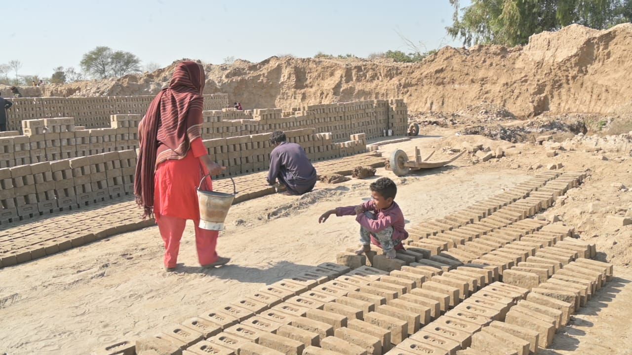 People working with bricks in an outdoor setting. Rows of bricks are drying in the sun, with a person and child nearby.