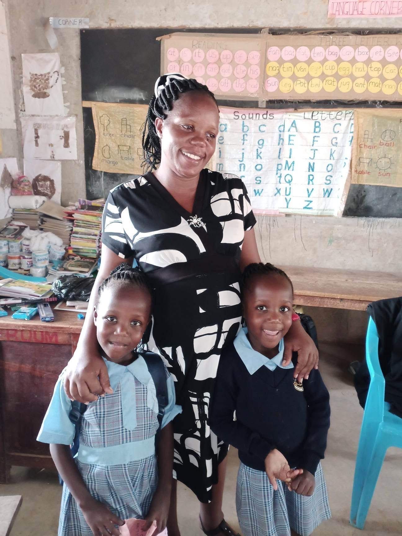 Woman with two children in a classroom, smiling. Children wear uniforms. Chalkboard and alphabet chart in background.