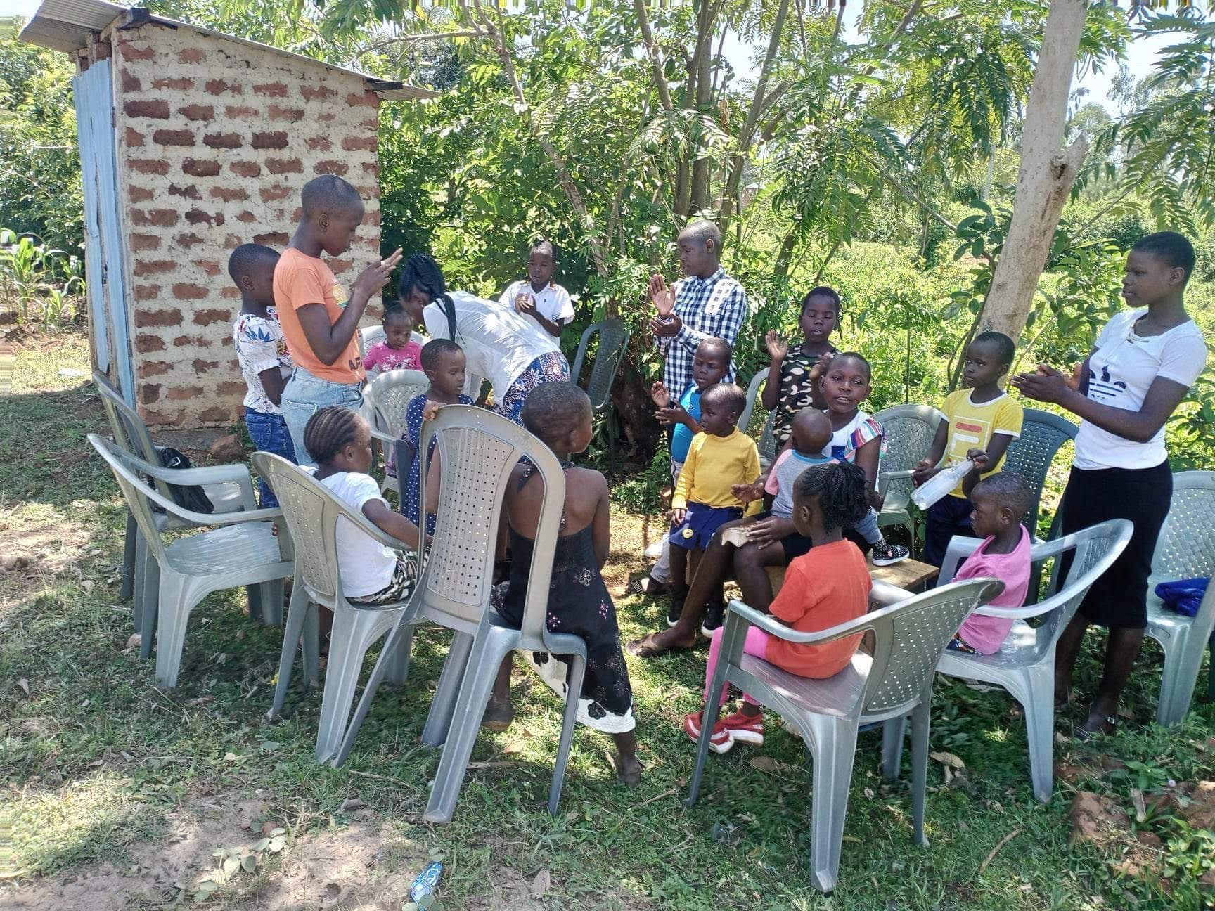 Children sitting in a circle on plastic chairs, listening to two adults in a sunny outdoor setting.
