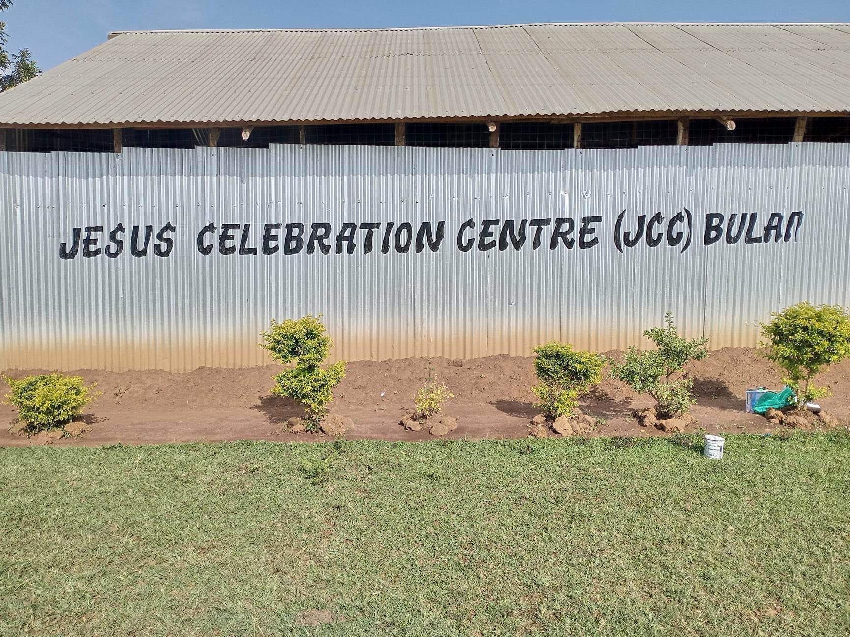 Jesus Celebration Centre (JCC) Bular, sign on corrugated metal wall, small bushes in front, green grass.