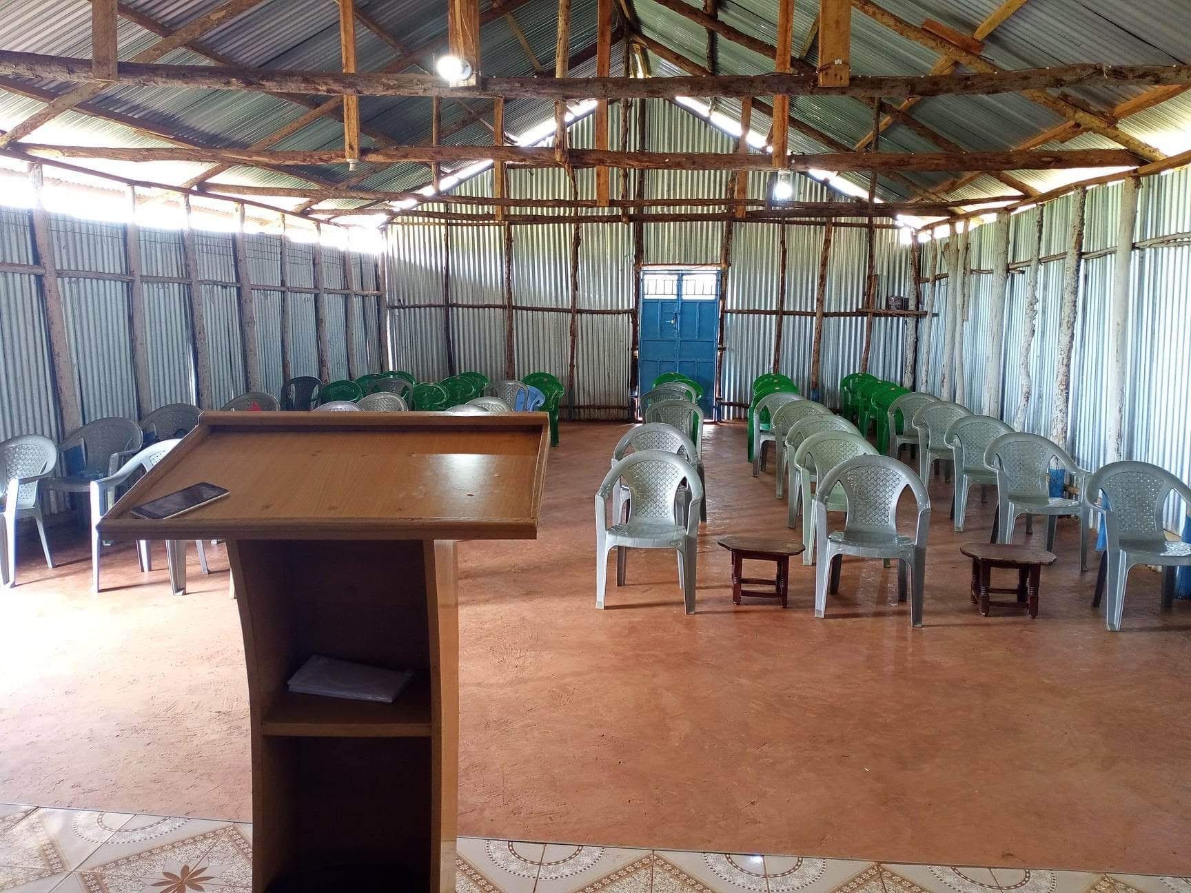 Interior of a simple building with rows of chairs, a wooden podium, and corrugated metal walls and roof.