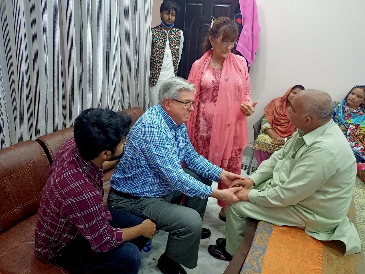 People in a room, two men hold hands. Others observe, a woman in pink, a young person in formal wear.
