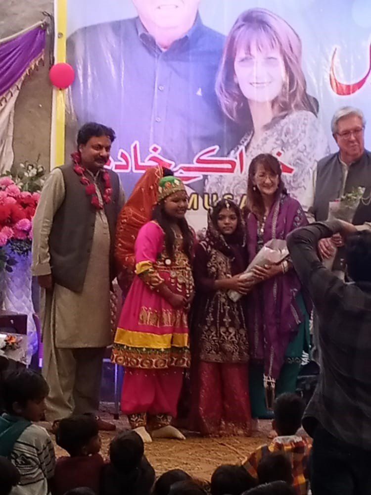 People on a stage presenting flowers to two girls in traditional dress. Event backdrop includes faces and text.