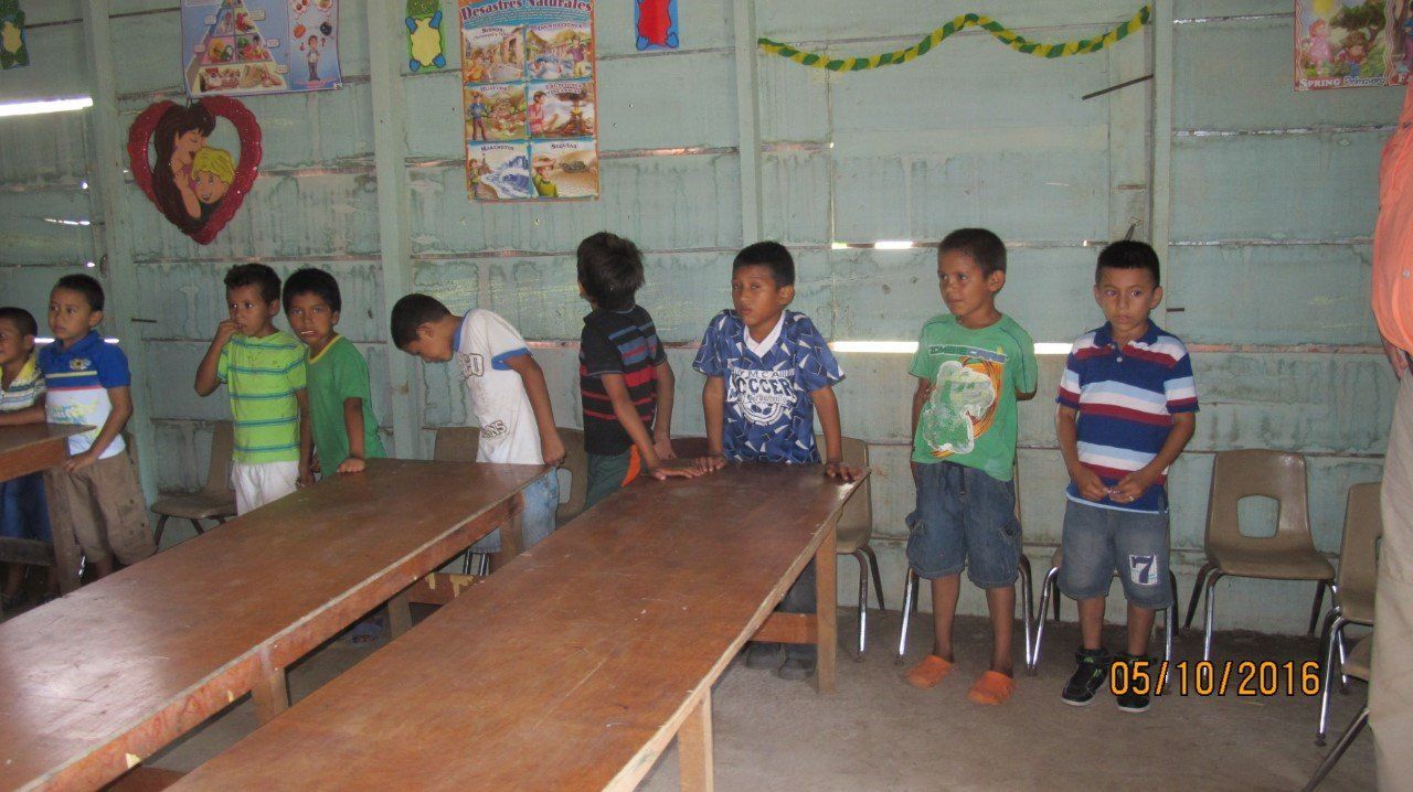 Children standing at desks in a classroom, some looking at the camera. Wooden tables and chairs.