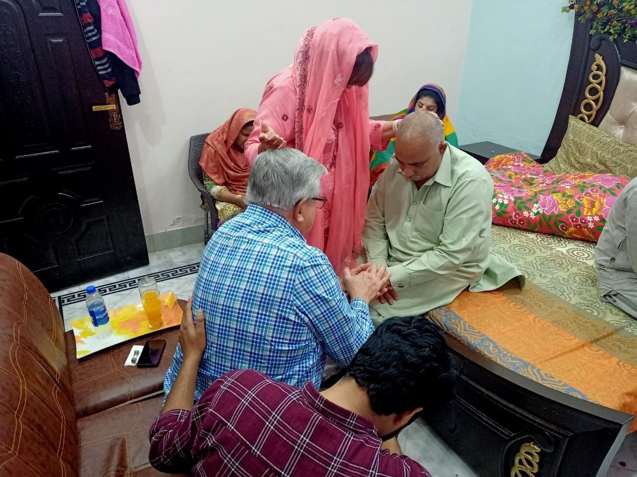 A group of people in a room; a person in a pink hijab is touching a man's head, another man is touching his hand.