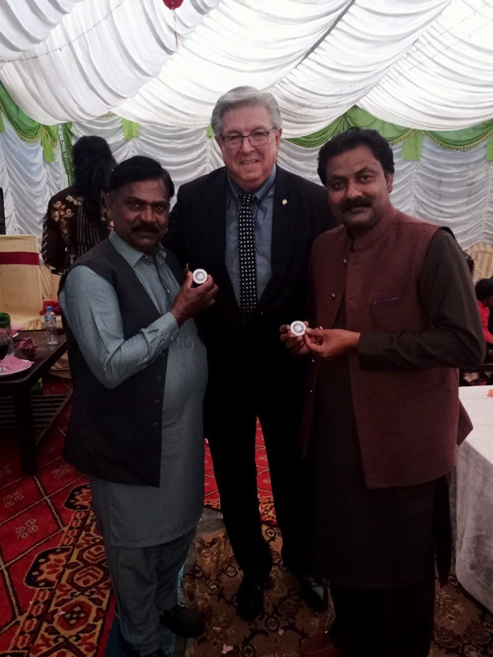 Three men holding objects, standing indoors with draped ceiling. Two men in traditional vests, one in a suit.