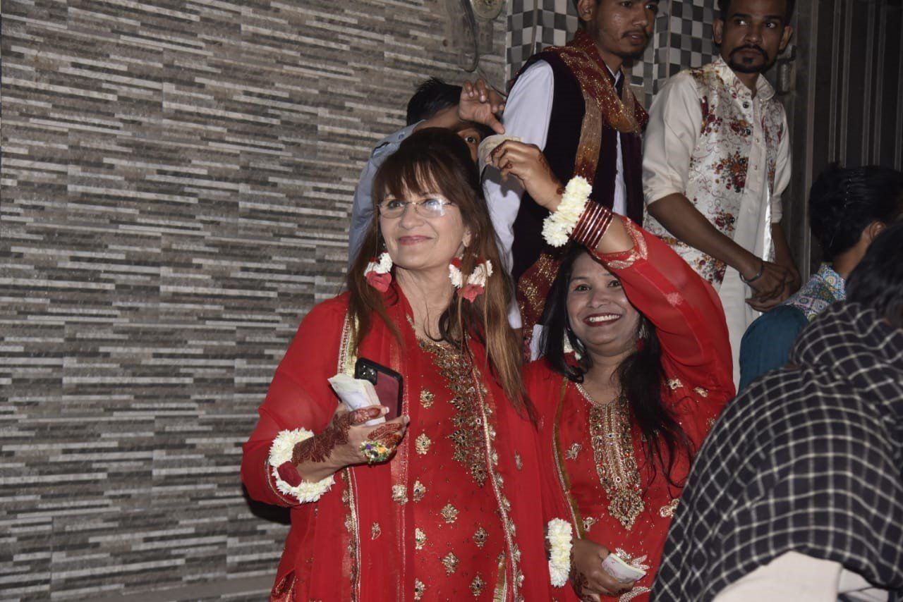 Two women in red embroidered outfits with floral garlands, smiling at an event; people in background.