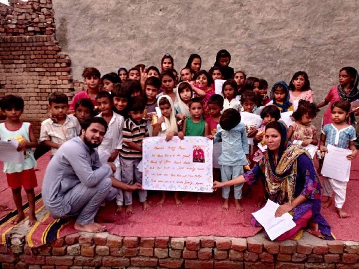 Group of children with two adults holding a sign, possibly for a cause, in a brick-walled setting.