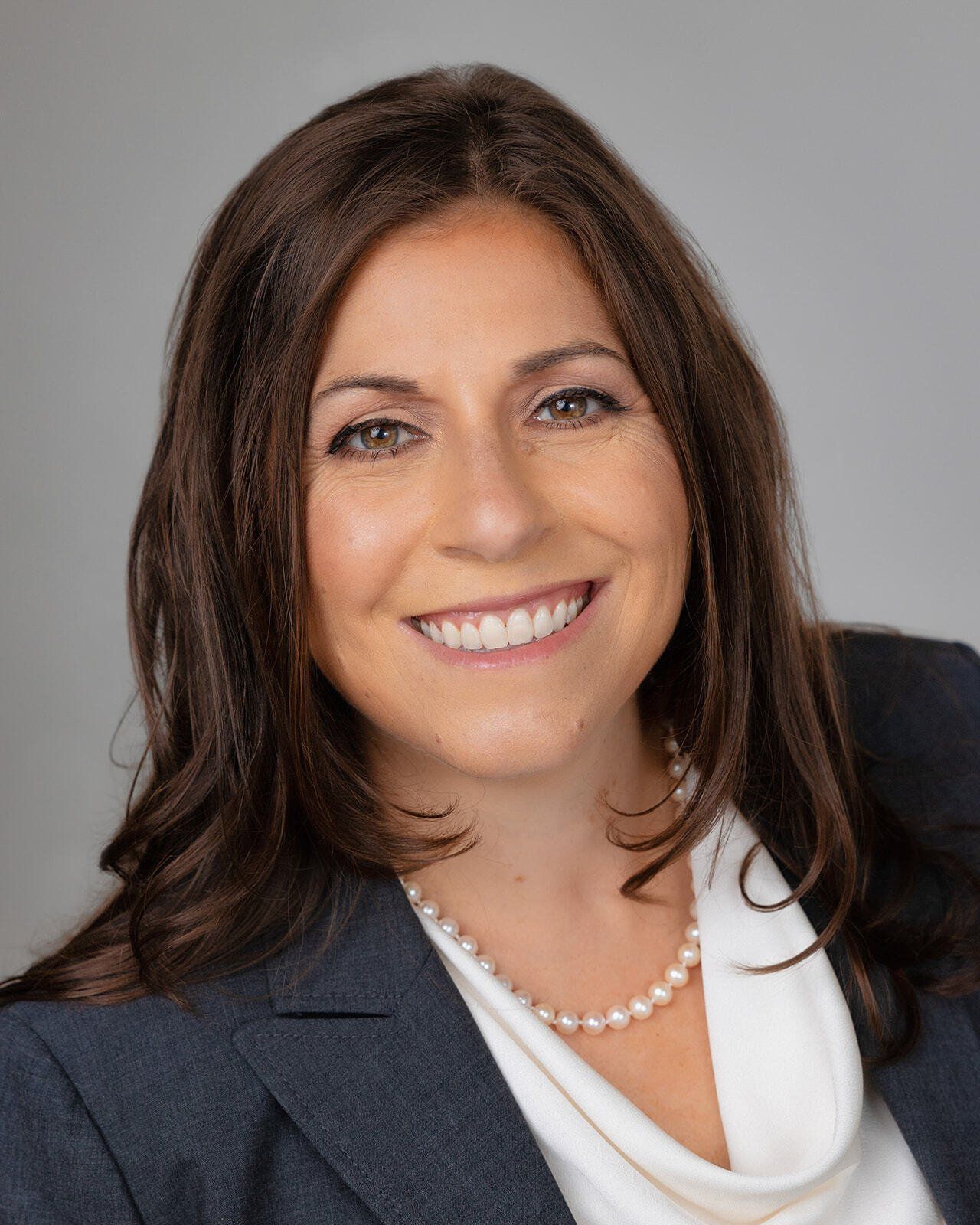 Professional headshot of a smiling person in a dark blazer and white blouse on a gray background