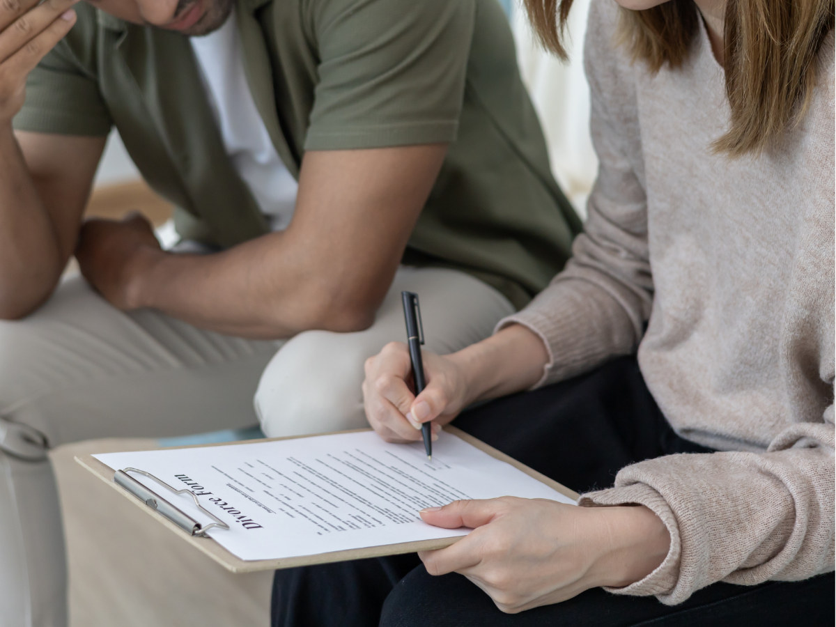 Two people sit on a sofa, one writing notes on a clipboard in a casual meeting setting.