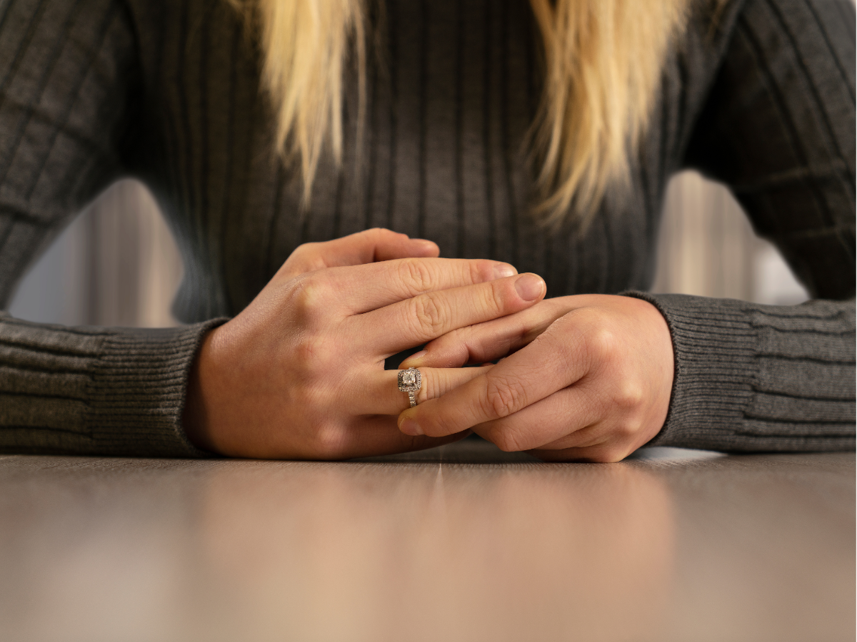 Hands clasped on a table, wearing a dark sweater and ring.