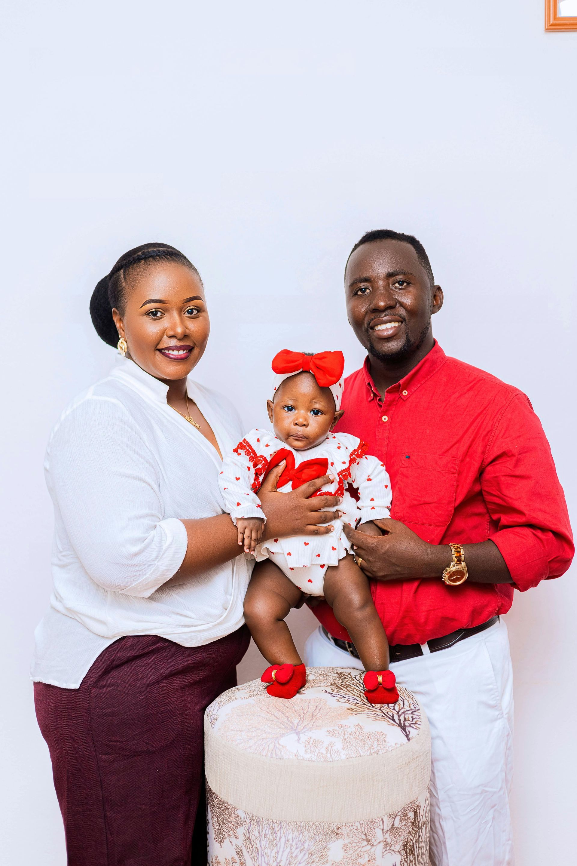 Family portrait with parents holding a baby in red, seated on a white cake prop against a light background