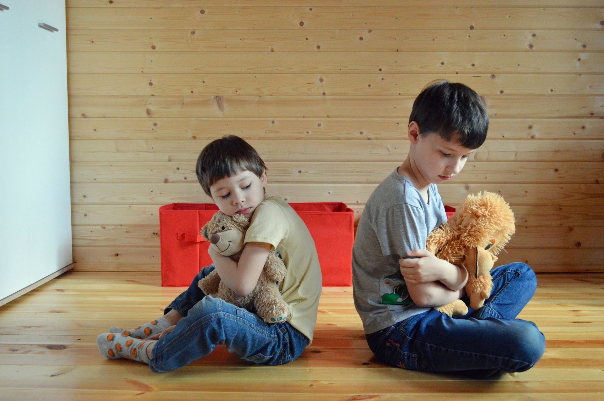 Two children sitting back-to-back on a wooden floor, each holding a stuffed teddy bear, with red blocks behind them