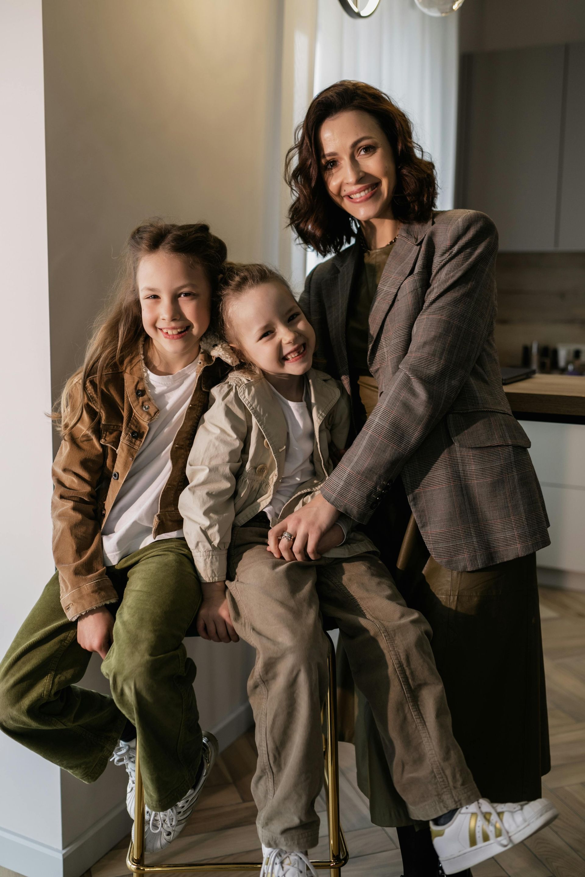 Three people sit on a kitchen counter, smiling in neutral-toned outfits.