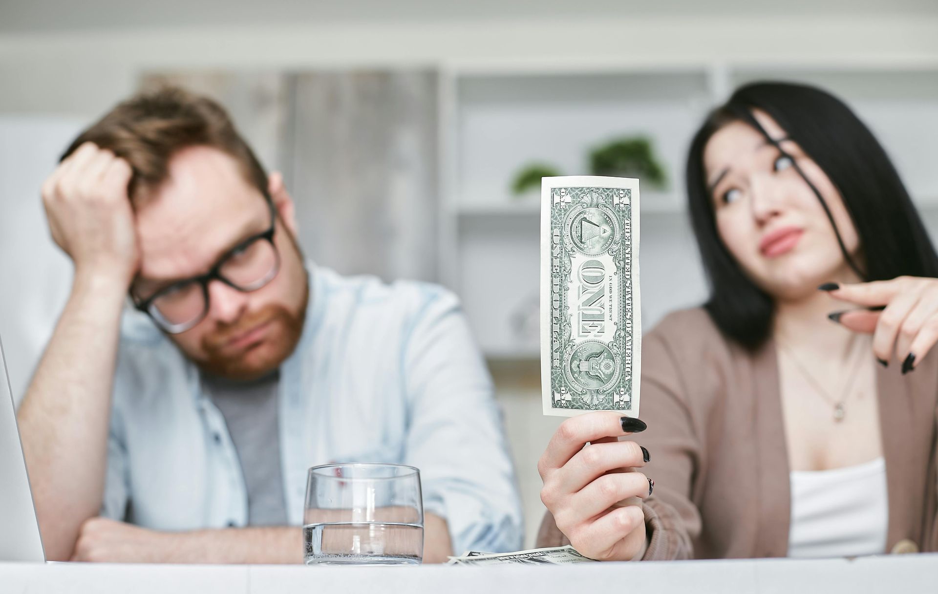 Two people at a table, one holding up a cash bill while the other looks frustrated