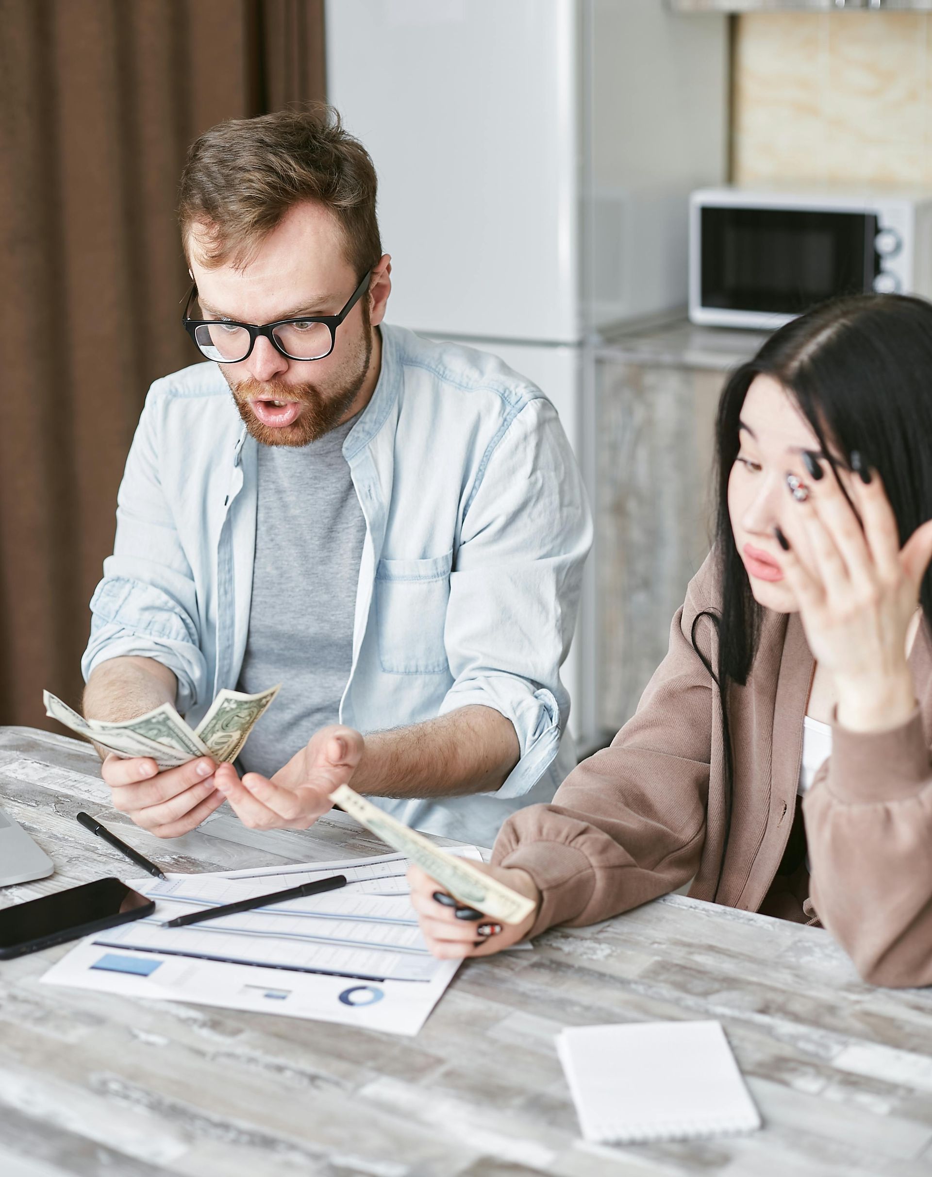 Two people at a table reviewing bills and money in a bright kitchen, one looking stressed.