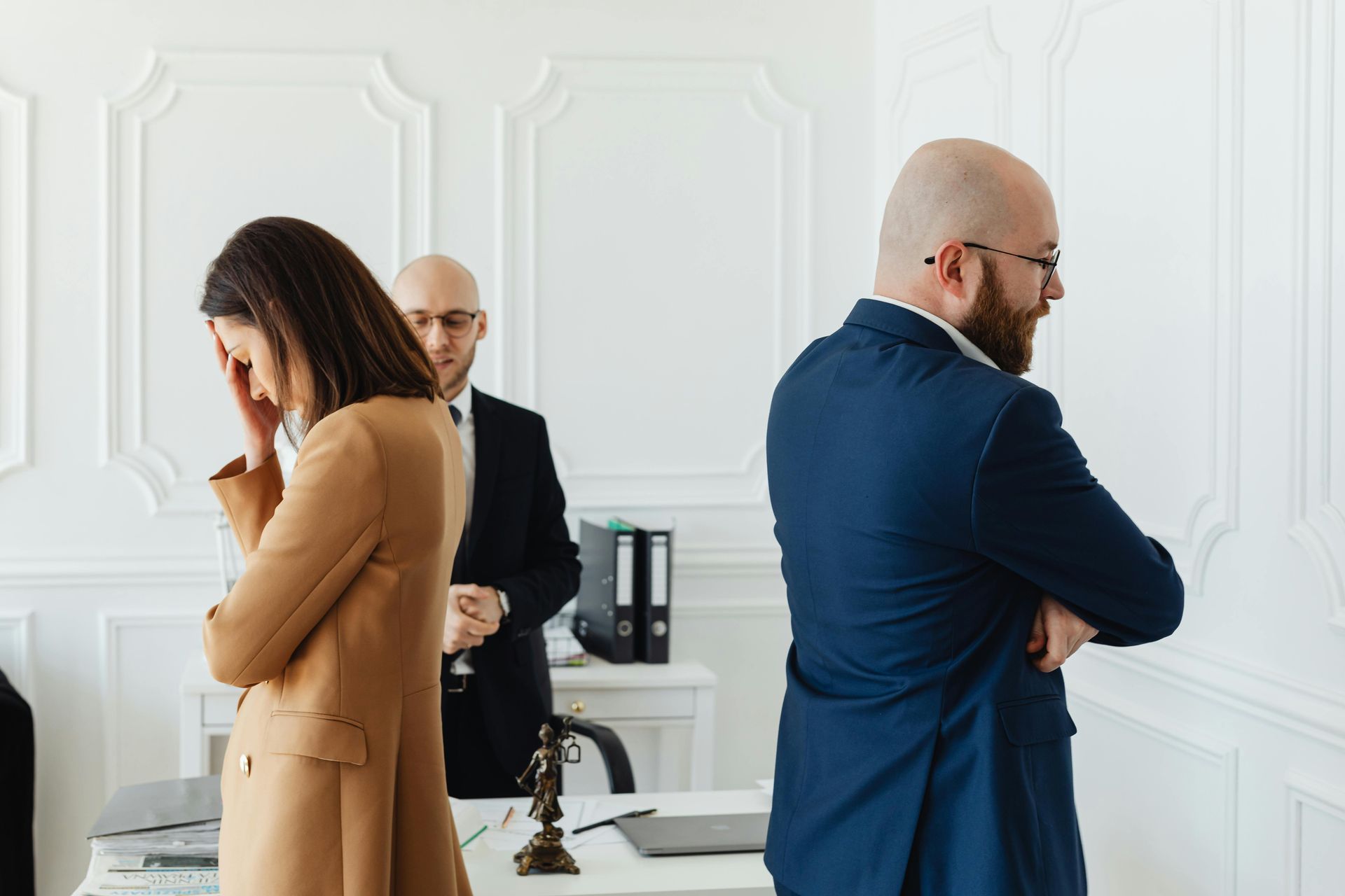 Three people in a bright office stand apart, looking serious near a desk.
