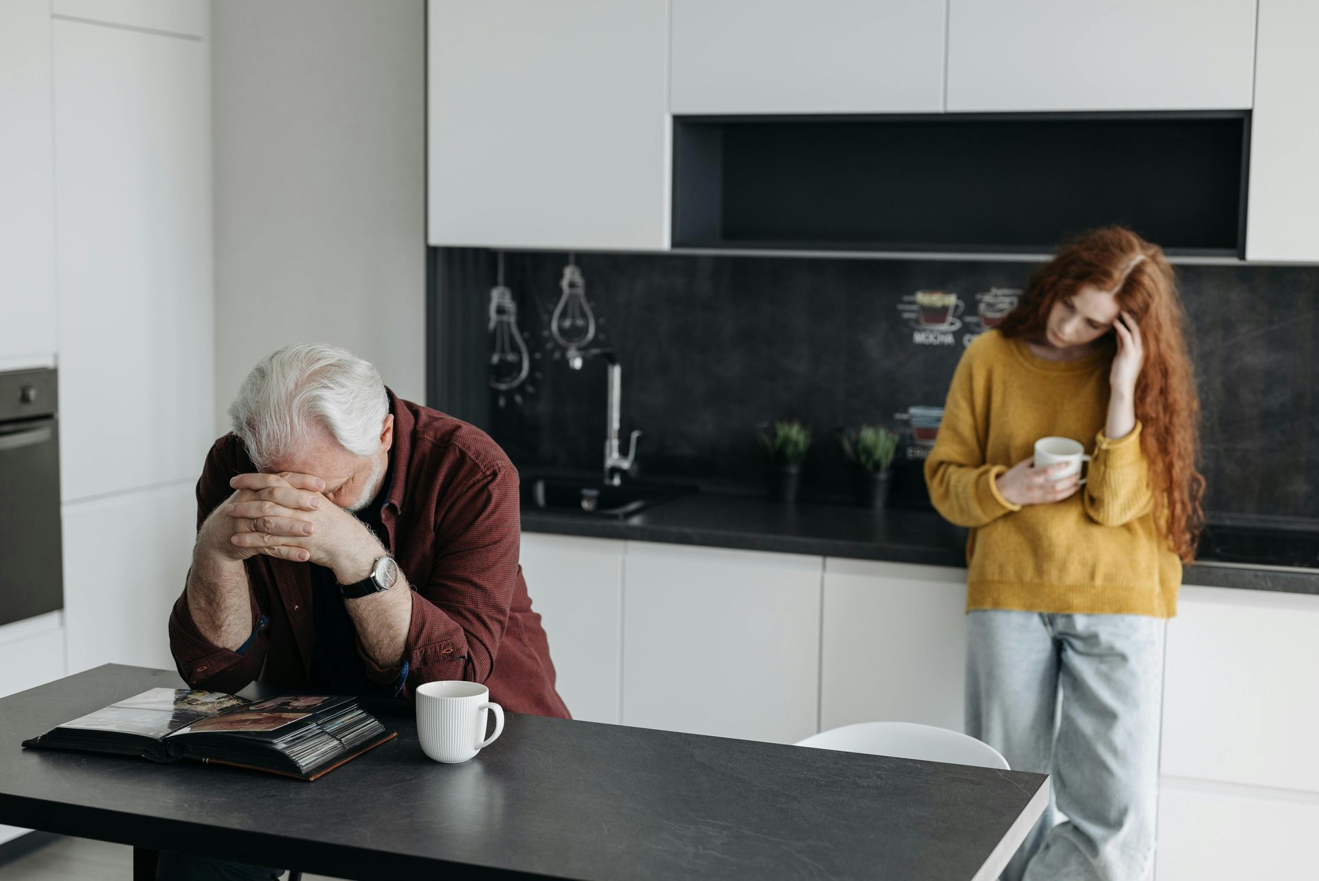 Person sitting at a kitchen table with head in hands while another stands nearby holding a mug
