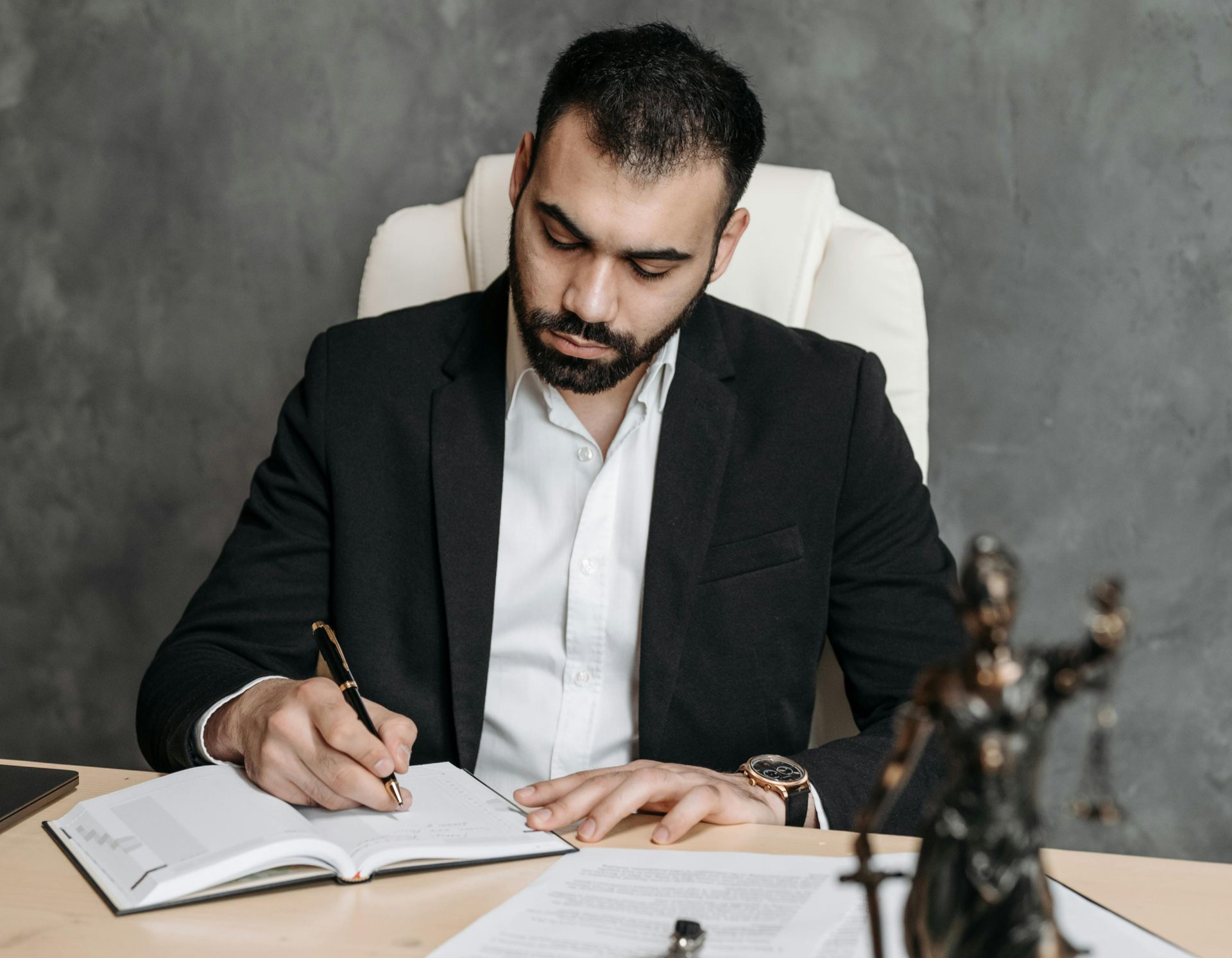 Man in a black suit writing in an open notebook at a desk, with a gray backdrop