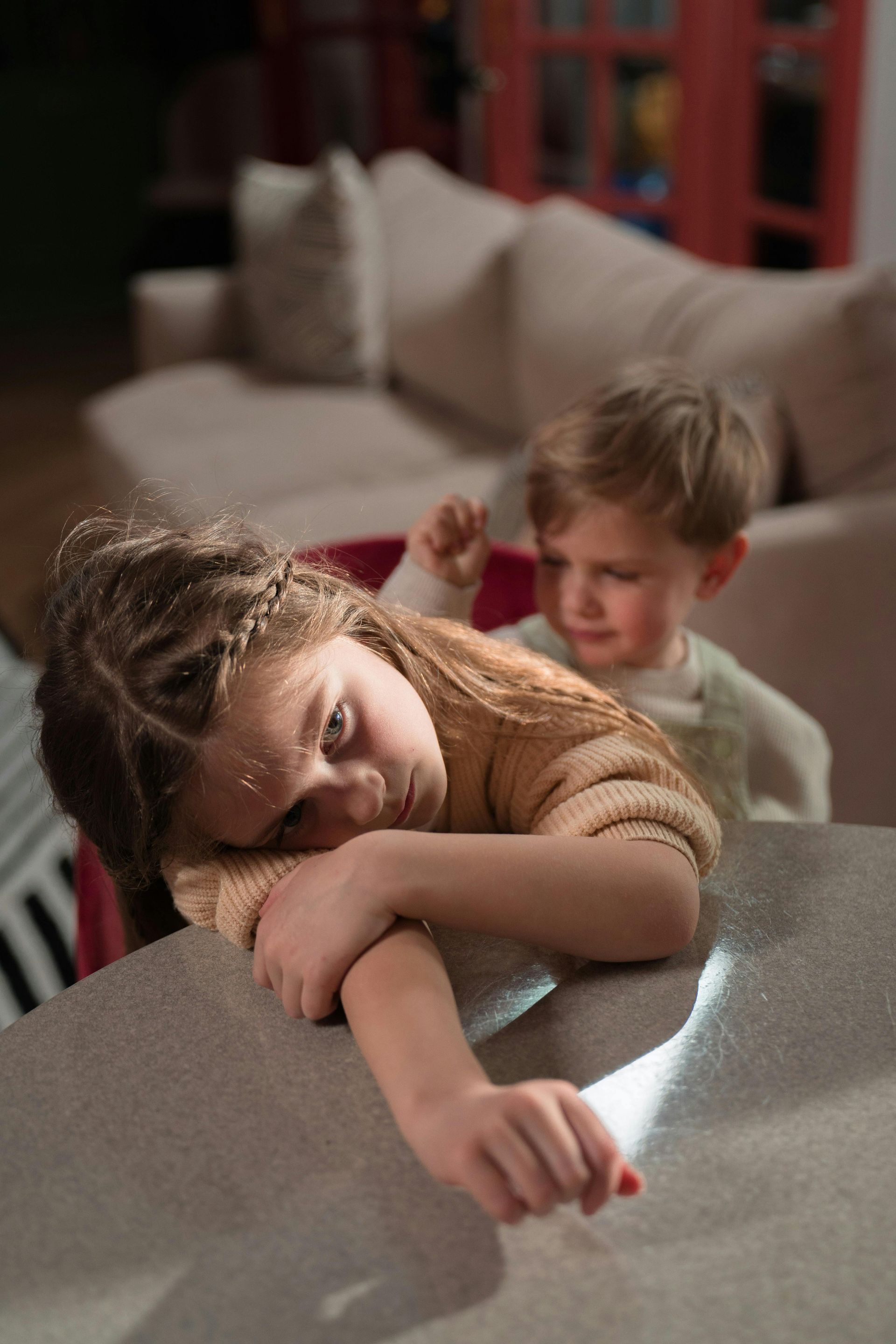 Two children resting on a couch, one with head on the table and arm outstretched, in a softly lit living room.