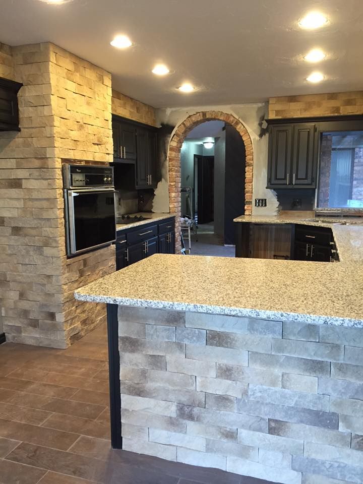 Modern kitchen with stone island, dark cabinets, built-in oven, and arched doorway under recessed lights