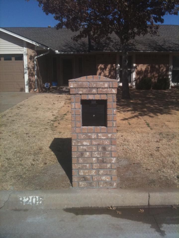 Brick mailbox on a suburban curb in front of a house, with a tree and driveway in the background