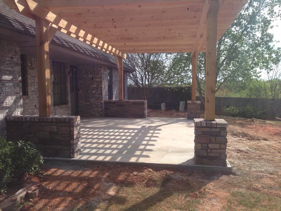 Covered stone patio with wooden beams, brick columns, and sunlight casting shadows on the concrete floor.
