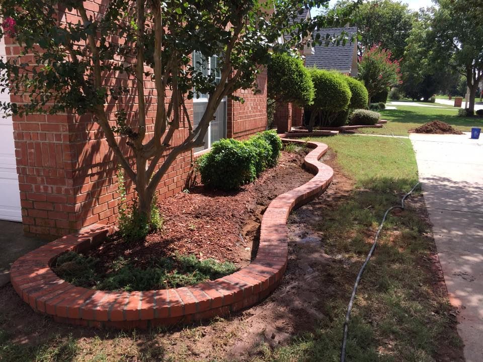 Curved brick garden border beside a house, with shrubs and a sidewalk on a sunny day