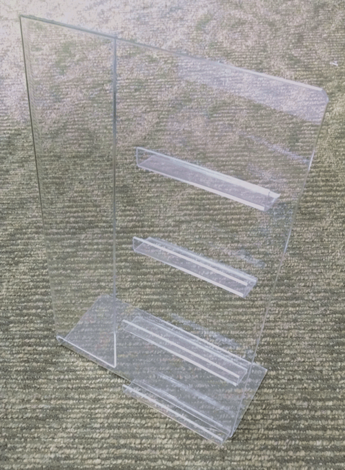 Clear acrylic brochure holder with three shelves on a textured carpeted floor