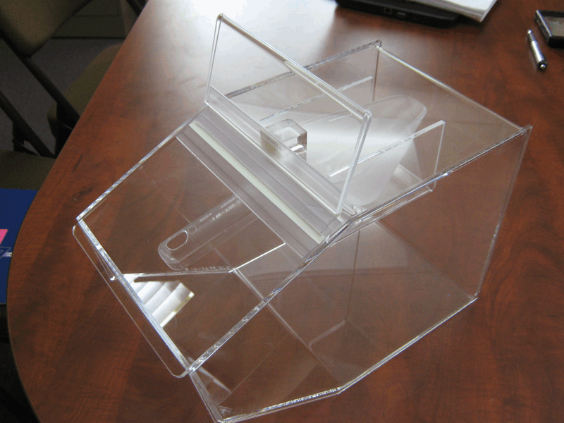 Clear acrylic ballot box on a wooden table, with a transparent hinged lid open.