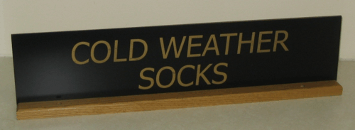 Black sign with gold text reading “COLD WEATHER SOCKS” on a wooden shelf￼