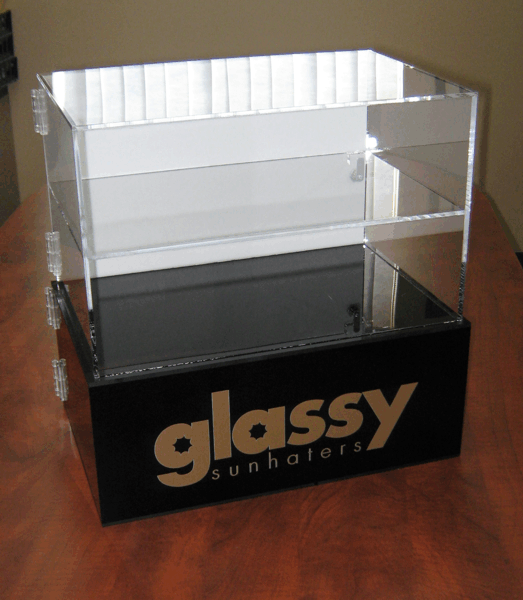 Clear acrylic display case with black base labeled “glassy sunglasses” on a wooden table