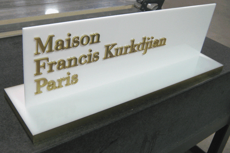 Gold “Maison Francis Kurkdjian Paris” sign on a white display stand.