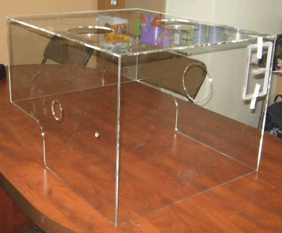 Clear acrylic box on a wooden table with a circular opening and side handle.