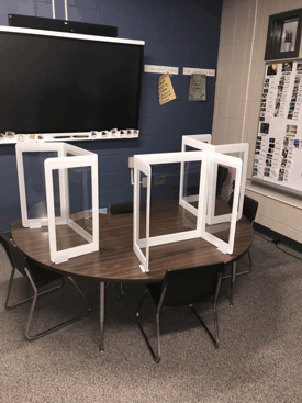 Classroom table with three white frame structures and black chairs around it on a gray carpet.