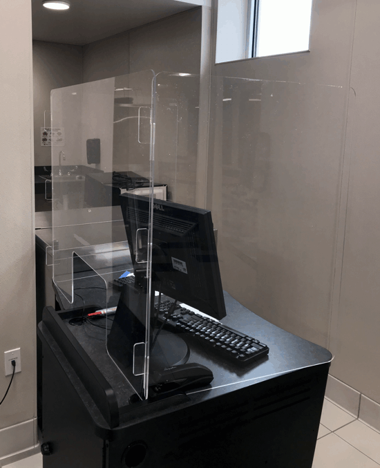 Empty service counter with plexiglass barrier, keyboard, and monitor in a tiled office or reception area.