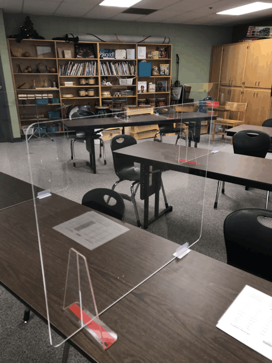 Classroom with desks, chairs, shelves, and clear plastic partitions on tables.