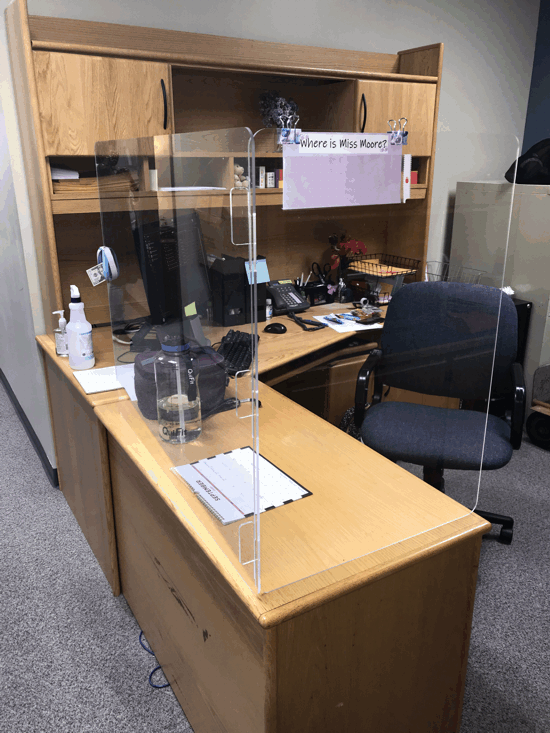 Office cubicle with wooden desk, clear plexiglass barriers, computer monitor, and chair, with a handwritten sign above.