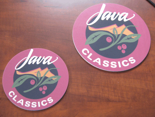 Two round pink Java Classics stickers with a bird and berries on a wooden surface.