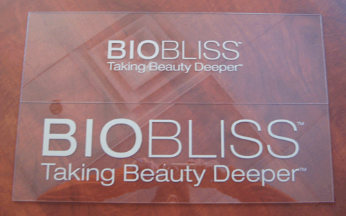 Two transparent BIOBLISS logo cards on a pink fabric background, reading “Taking Beauty Deeper.”