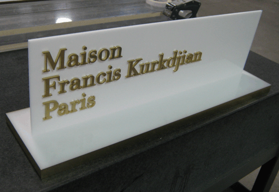 White acrylic display sign reading “Maison Francis Kurkdjian Paris” in gold letters on a black counter