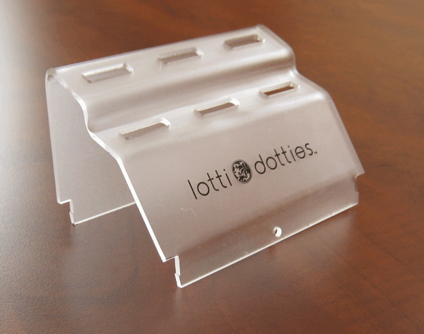 Clear acrylic display stand with slotted grooves and “lotti dotties” logo on a wooden surface