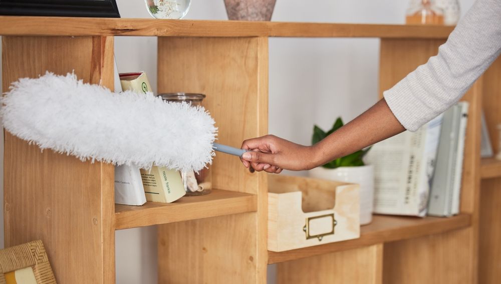 Hand dusting a wooden shelf with a white duster near books and storage boxes