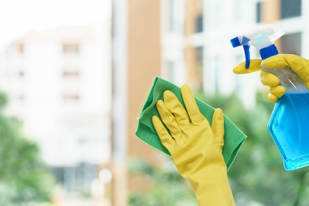 Person Wearing Yellow Gloves Spraying Blue Cleaner on a Window — Crystal Clear In Glen Innes, NSW